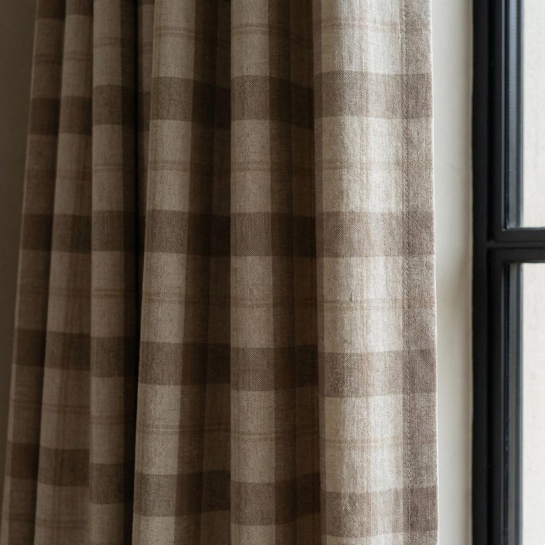 May include: Close-up of a brown and beige plaid curtain panel. The fabric has a textured appearance and is hanging near a window with a black frame. The curtain is made of a woven material.