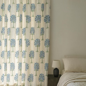 Op de afbeelding: Crèmekleurig gordijn met een herhaald patroon van blauwe en groene gestileerde bomen. Het gordijn hangt voor een bed met een crèmekleurig dekbed en kussen. Een kleine lamp staat op een donker nachtkastje.