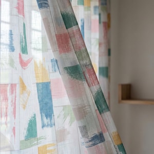 May include: Sheer white curtains with a colorful abstract brushstroke pattern. The design features rectangular and irregular shapes in shades of pink, blue, green, yellow, and peach. The curtains are hanging in front of a window.