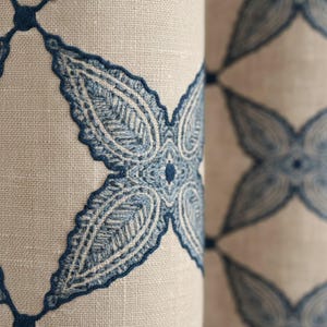 以下が含まれることがあります： 繰り返しの刺繍模様が施された生地のクローズアップ。デザインは、ニュートラルなベージュの背景に青い花のモチーフが特徴です。刺繍は詳細で質感のある外観で、装飾的なテキスタイルを作成しています。