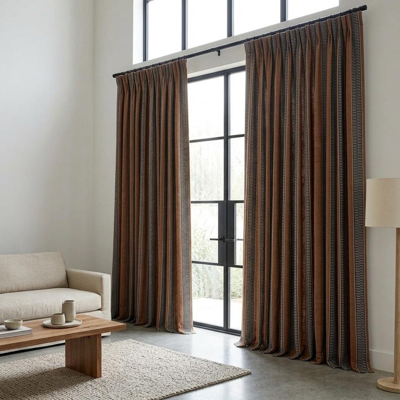 Pu&ograve; includere: Tende plissettate dal pavimento al soffitto in un ricco color marrone ruggine, caratterizzate da strisce verticali con sottili motivi bianchi e neri. Le tende sono appese a un bastone in metallo nero davanti a una grande finestra con telaio nero.