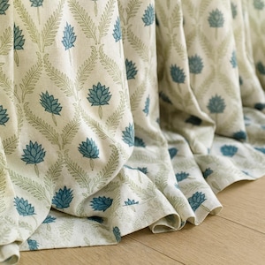 Op de afbeelding: Crèmekleurige stof met een herhalend patroon van blauwe lotusbloemen en groene bladeren. De stof is gedrapeerd, wat de textuur en de beweging van het materiaal laat zien. Het ontwerp is een mix van bloem- en geometrische elementen.