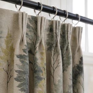 May include: Beige curtain panels with a forest-themed print. The design features stylized trees in shades of green and gray, with delicate black branches. The curtains hang from a black rod with silver rings, creating a natural, serene aesthetic.