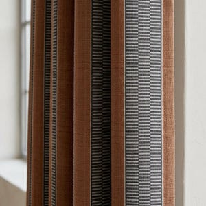 Pu&ograve; includere: Una tenda plissettata caratterizzata da pannelli verticali in tessuto color arancio bruciato a tinta unita, alternati a strisce pi&ugrave; strette con motivi geometrici grigio scuro e bianchi. Il tessuto ha una trama visibile e cade in pieghe.