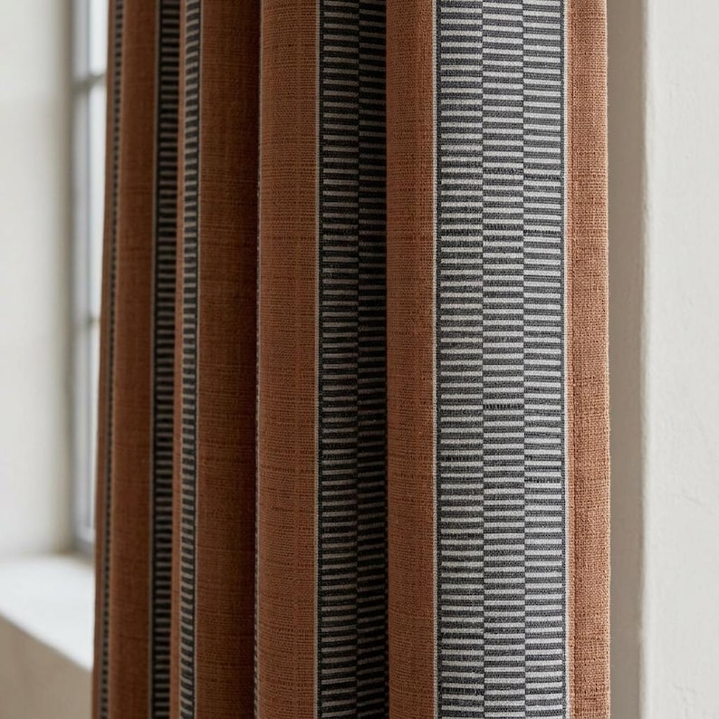 Pu&ograve; includere: Una tenda plissettata caratterizzata da pannelli verticali in tessuto color arancio bruciato a tinta unita, alternati a strisce pi&ugrave; strette con motivi geometrici grigio scuro e bianchi. Il tessuto ha una trama visibile e cade in pieghe.