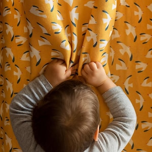 Op de afbeelding: Handen van een kind grijpen een mosterdgele gordijn met een herhaald patroon van witte vogels met blauwe accenten. Het kind draagt een grijs shirt met lange mouwen. Het gordijn is het middelpunt.