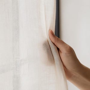 Könnte beinhalten: Nahaufnahme eines transparenten, cremefarbenen Vorhangs. Der Stoff wird von einer Hand gehalten, wodurch die Textur und das Gewebe des Materials sichtbar werden. Der Vorhang scheint aus einem leichten, atmungsaktiven Stoff zu bestehen, der zum Filtern von Licht geeignet ist.