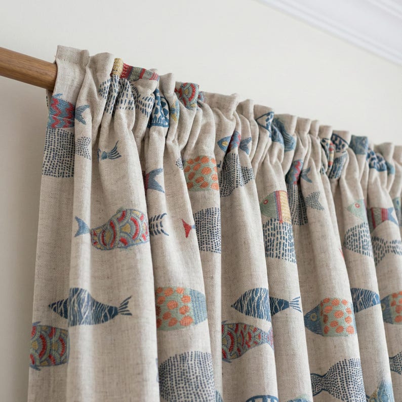 May include: Beige linen curtains with a repeating pattern of colorful fish in blue, orange, and red. The curtains are gathered at the top and hang in soft folds. The fish design adds a nautical theme to the room.