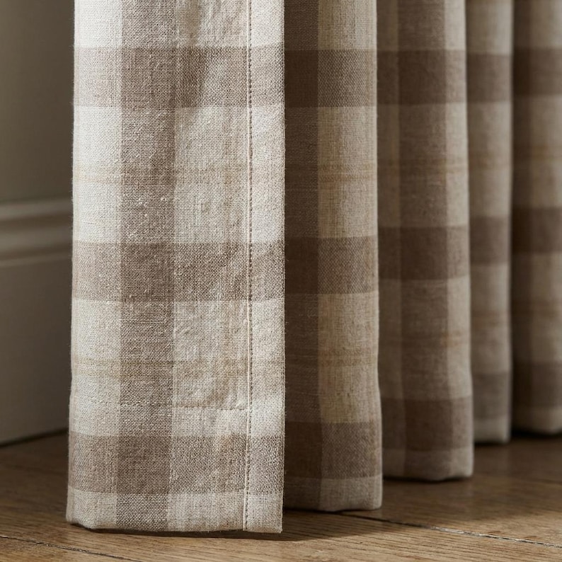 May include: Close-up of beige and brown checkered curtains. The fabric has a textured appearance, with visible threads and a subtle sheen. The curtains are hanging vertically, with a visible seam down the side.