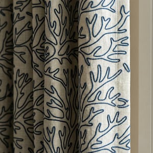 Könnte beinhalten: Nahaufnahme von beigefarbenen Vorhängen mit einem sich wiederholenden Muster aus dunkelblauen, korallenartigen Designs. Der Stoff ist strukturiert, und die Vorhänge sind teilweise geschlossen, wodurch eine weiße Wand im Hintergrund sichtbar wird. Das Design erinnert an Meereslebewesen.