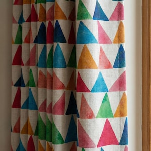 May include: Curtains with a repeating pattern of colorful triangles in shades of red, blue, green, and yellow on a neutral background. The fabric is textured and appears to be linen or a similar material. The curtains are hanging in front of a window.