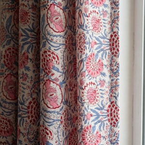 May include: Floral patterned curtains with a repeating design of pink and red flowers, blue leaves, and stems on a white background. The fabric is folded in vertical pleats, creating a textured appearance. The curtains are near a window.