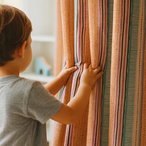 Könnte beinhalten: Hände eines Kindes ziehen einen gemusterten Vorhang zurück. Der Vorhang hat vertikale Streifen in Orange-, Türkis-, Rosa- und Cremetönen. Der Stoff hat ein strukturiertes Aussehen und verleiht der Fensterdekoration visuelles Interesse.