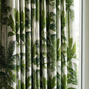 Op de afbeelding: Gordijnen met een witte achtergrond en een groene tropische bladerprint. Het ontwerp heeft een herhalend patroon van palmbladeren en ander tropisch gebladerte in verschillende groentinten. De gordijnen hangen voor een raam.