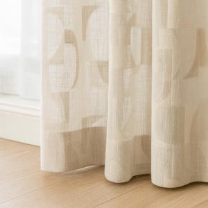 Könnte beinhalten: Beige Leinen-Vorhänge mit geometrischem Muster. Die Vorhänge sind teilweise zugezogen und zeigen ein Fenster und eine hellfarbige Wand. Der Stoff hat eine strukturierte Optik, und die Vorhänge hängen in weichen Falten.