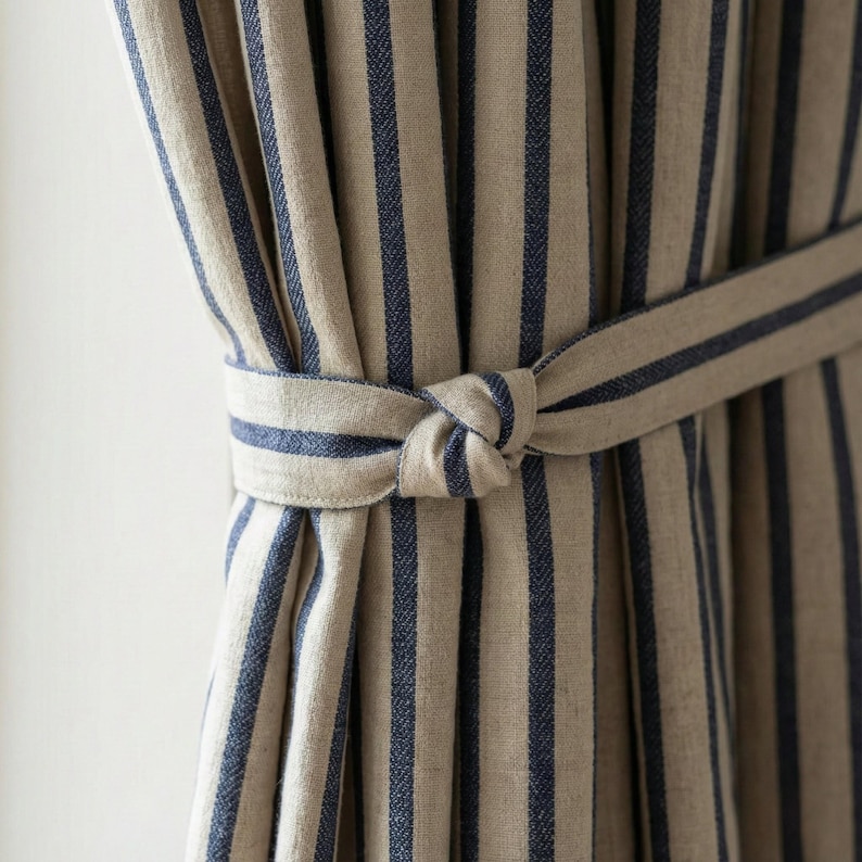 Op de afbeelding: Close-up van beige en marineblauwe gestreepte gordijnen, vastgebonden met een bijpassende stoffen riem. De gordijnen hebben verticale strepen en zijn gemaakt van een textuurstof. De riem is in een knoop gelegd.
