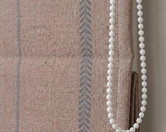 May include: A beige fabric roman shade featuring a vertical blue herringbone pattern stripe and a white plastic beaded chain mechanism for operation. The textured material displays a woven weave with visible horizontal folds and stitched seams.