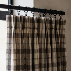 Peut inclure: Gros plan sur un rideau à carreaux marron et beige. Le rideau est suspendu à une tringle noire avec des anneaux et des pinces. Le tissu est plissé, créant un effet texturé. Le rideau est en tissu épais.
