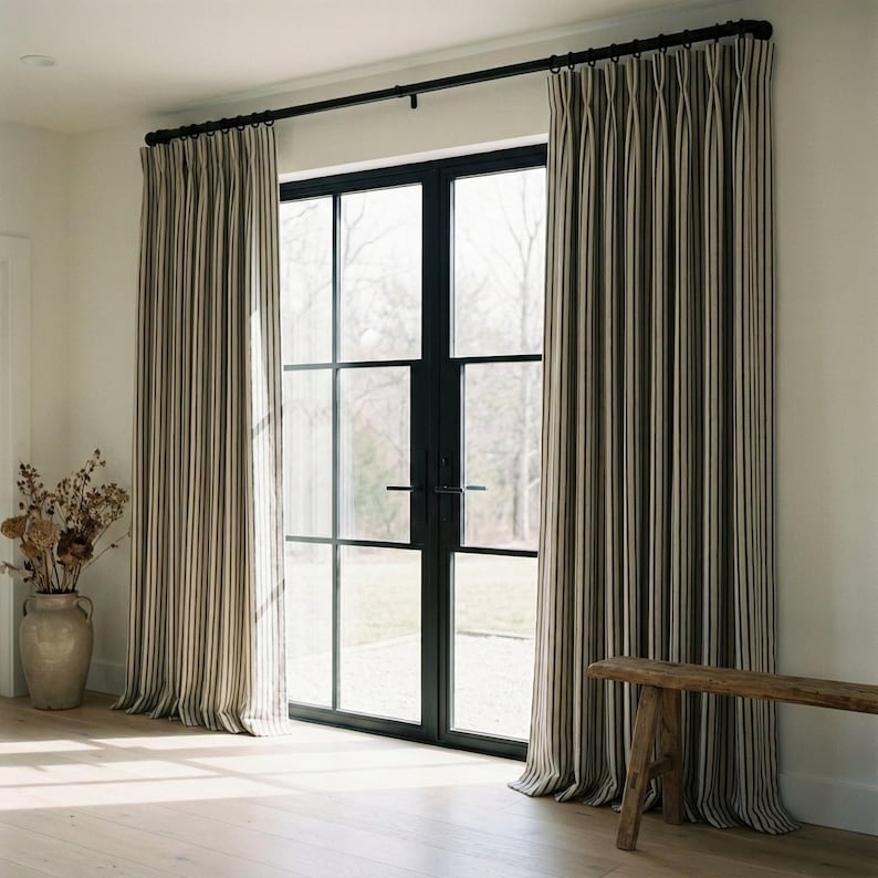 Op de afbeelding: Gestreepte gordijnen in neutrale tinten omlijsten een groot raam met zwart frame. De gordijnen hebben verticale strepen in beige, bruin en wit. Onder het raam staat een houten bank en links staat een vaas met droogbloemen.
