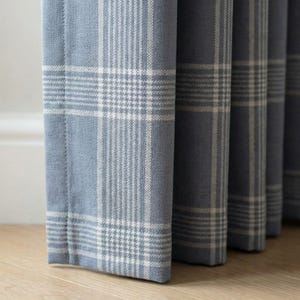 May include: Close-up of blue plaid curtains with a white grid pattern. The fabric has a soft texture and is hanging in vertical folds. The curtains are a light blue color.