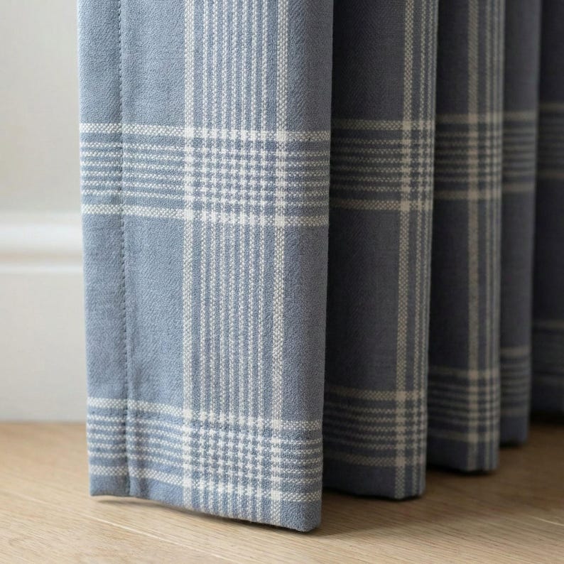 May include: Close-up of blue plaid curtains with a white grid pattern. The fabric has a soft texture and is hanging in vertical folds. The curtains are a light blue color.