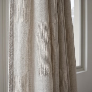 May include: Beige linen curtain with vertical textured stripes and rectangular patterns. The curtain is partially drawn, revealing a glimpse of a window. The fabric has a natural, slightly wrinkled appearance.