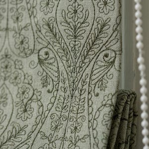 Può includere: Un pannello di tessuto verde salvia chiaro con motivi floreali e paisley ricamati in verde scuro. Il tessuto presenta una trama visibile ed è mostrato con una sezione piegata, che mostra un design decorativo coerente su tutta la superficie.