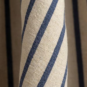 Op de afbeelding: Close-up van opgerolde stof met een natuurlijke beige achtergrond en opvallende, diagonale marineblauwe strepen. De stof heeft een getextureerde uitstraling, wat wijst op linnen of een vergelijkbaar materiaal. De strepen zijn gelijkmatig verdeeld en cre&euml;ren een klassieke, nautische esthetiek.