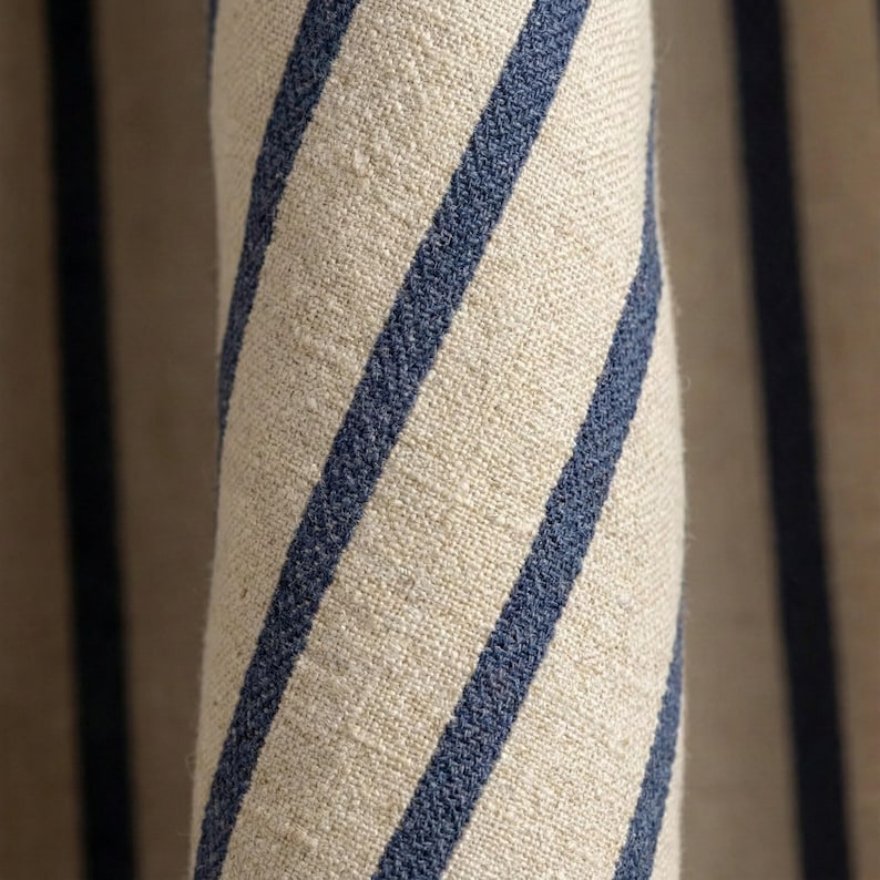 Op de afbeelding: Close-up van opgerolde stof met een natuurlijke beige achtergrond en opvallende, diagonale marineblauwe strepen. De stof heeft een getextureerde uitstraling, wat wijst op linnen of een vergelijkbaar materiaal. De strepen zijn gelijkmatig verdeeld en cre&euml;ren een klassieke, nautische esthetiek.