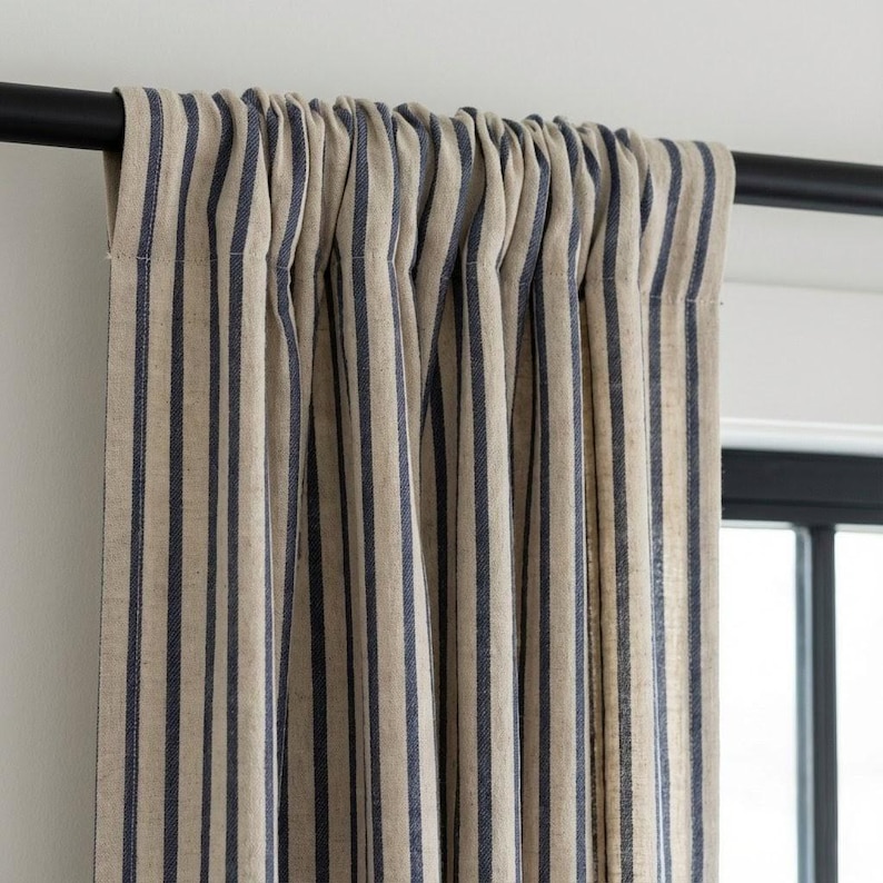 Op de afbeelding: Beige en marineblauwe gestreepte gordijnpanelen die aan een zwarte stang hangen. De gordijnen hebben verticale strepen en zijn gemaakt van een textuurstof. De gordijnen bedekken gedeeltelijk een raam.