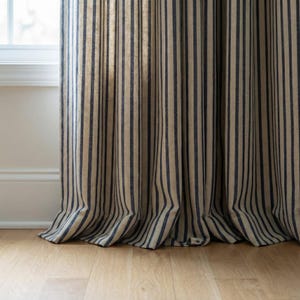 Op de afbeelding: Beige en marineblauwe gestreepte gordijn. De stof is gedrapeerd in verticale plooien, met de strepen van boven naar beneden. Het materiaal lijkt linnen of een vergelijkbare stof te zijn. Het gordijn staat naast een wit raamkozijn en staat op een lichte houten vloer.