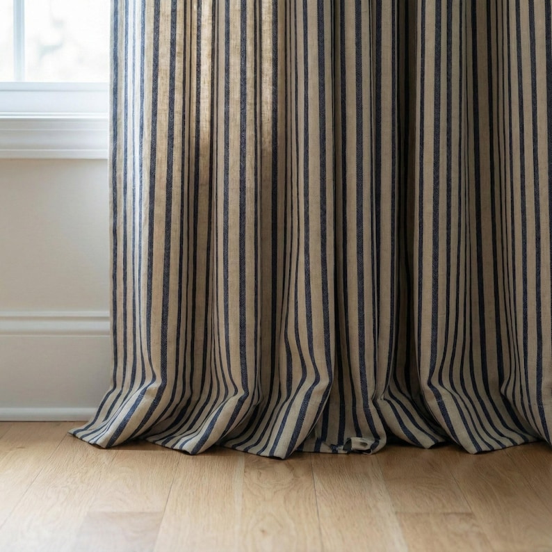Op de afbeelding: Beige en marineblauwe gestreepte gordijn. De stof is gedrapeerd in verticale plooien, met de strepen van boven naar beneden. Het materiaal lijkt linnen of een vergelijkbare stof te zijn. Het gordijn staat naast een wit raamkozijn en staat op een lichte houten vloer.
