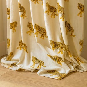 May include: Cream-colored curtain panels with a repeating pattern of yellow and black tiger illustrations. The fabric drapes in soft folds, showcasing the detailed tiger designs. The curtains are likely made of a lightweight material.