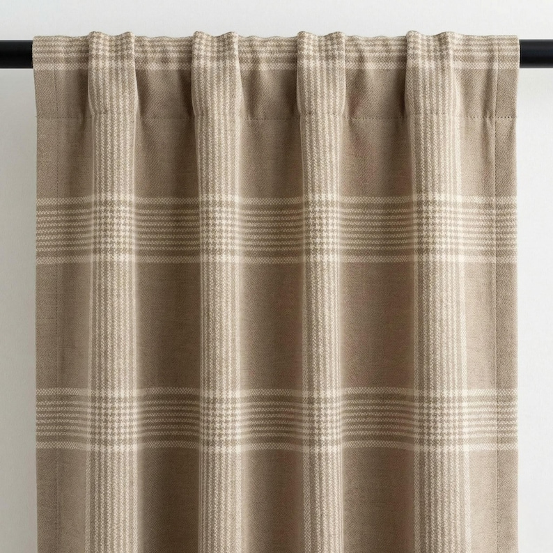 May include: Tan and cream plaid curtain panel. The curtain features a classic plaid pattern with horizontal and vertical lines. The fabric is a neutral color, suitable for various interior design styles. The curtain is hanging from a black rod.