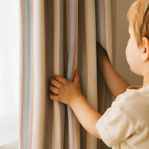 May include: Cream-coloured striped curtains with vertical red and blue stripes. A child's hands are pulling the curtain open. The curtains are made of a textured fabric.