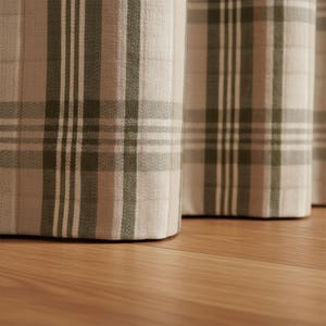 May include: Close-up of three fabric-covered ottomans with a plaid pattern in shades of beige, white, and olive green. The ottomans are positioned on a wooden floor, with the focus on the textured fabric and the plaid design.