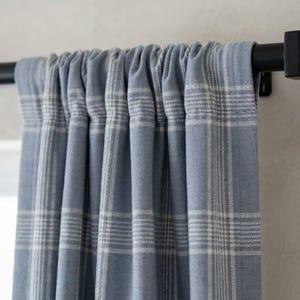 May include: Blue plaid curtains hanging from a black rod. The curtains have a light blue background with a white plaid pattern. The fabric is gathered at the top, creating soft folds. The curtains are a home decor item.
