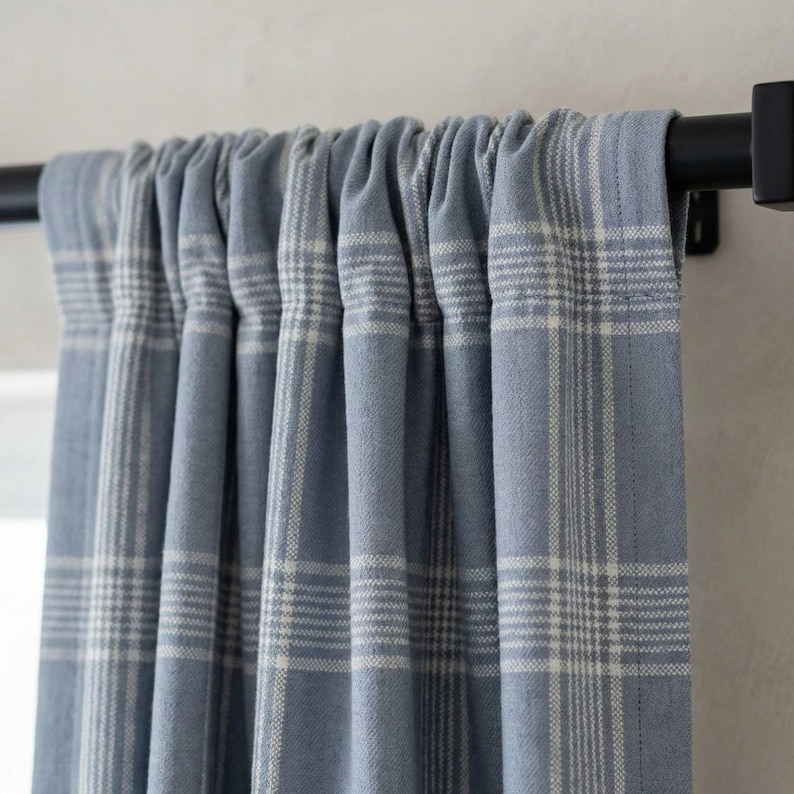 May include: Blue plaid curtains hanging from a black rod. The curtains have a light blue background with a white plaid pattern. The fabric is gathered at the top, creating soft folds. The curtains are a home decor item.