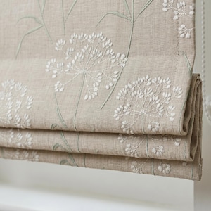 May include: A beige roman shade featuring white embroidered floral patterns and light green stems. The fabric has a visible woven texture and is folded into horizontal pleats. A white beaded pull chain is visible on the right side of the shade.
