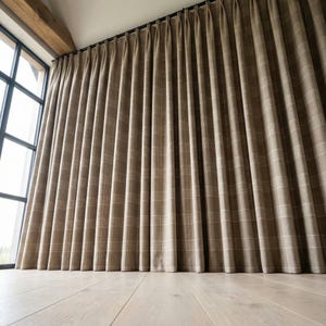 May include: Brown plaid curtains hanging from a black rod, covering a large window. The curtains have a subtle grid pattern in a lighter shade of brown. The floor is made of light-colored wood.