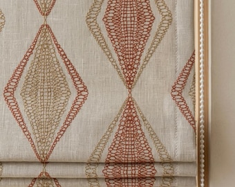 May include: A beige fabric roman shade featuring a repeating diamond pattern embroidered in rust orange and metallic gold thread. The design consists of large, interconnected geometric shapes with intricate, lace-like stitching details throughout.