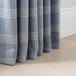 May include: Close-up of blue plaid curtains with white and gray accents. The fabric is folded in vertical pleats, revealing the pattern. The curtains are hanging in front of a white wall and light-colored wooden floor.