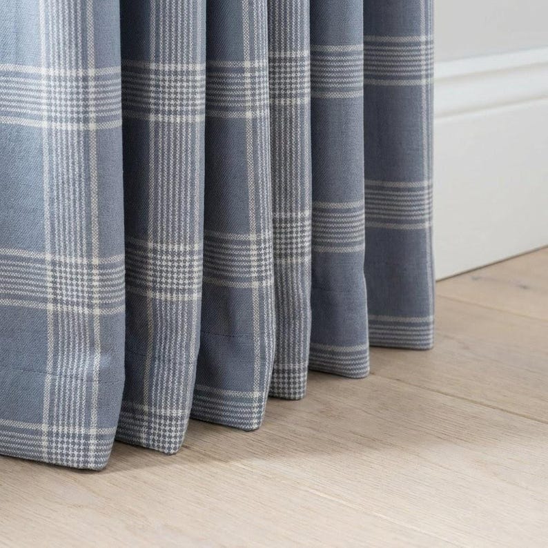 May include: Close-up of blue plaid curtains with white and gray accents. The fabric is folded in vertical pleats, revealing the pattern. The curtains are hanging in front of a white wall and light-colored wooden floor.