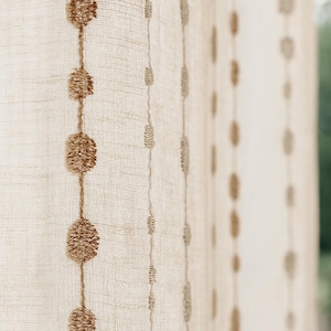 Può includere: Tenda in lino beige con file verticali di abbellimenti ovali strutturati in una tonalità marrone più scura. La tenda ha una trama sottile e tessuta, e il design aggiunge interesse visivo.