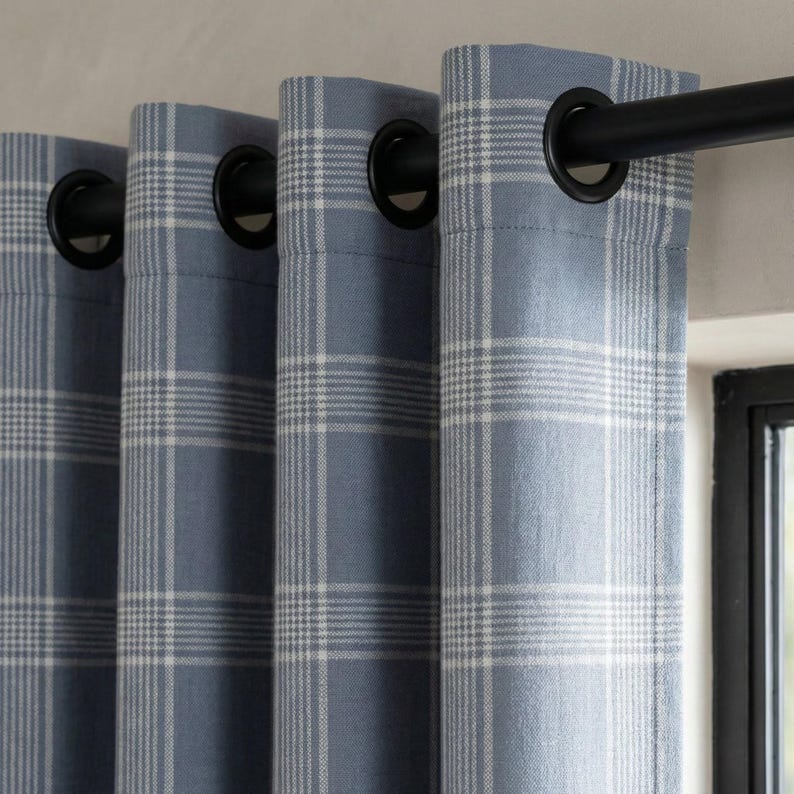 May include: Blue plaid curtains with black grommets hanging from a black curtain rod. The curtains have a light blue background with a white plaid pattern. The curtains are hanging in front of a window.