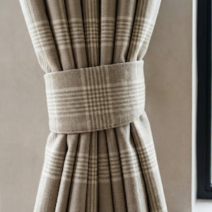 May include: Close-up of a beige plaid curtain held by a matching fabric tie. The curtain has a classic plaid pattern in light and dark beige tones. The tie is wrapped around the gathered fabric, creating a neat, decorative look. The curtain is hanging near a window.