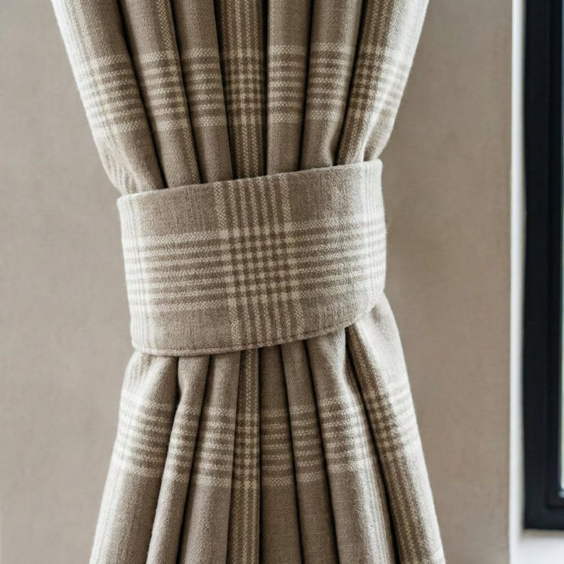 May include: Close-up of a beige plaid curtain held by a matching fabric tie. The curtain has a classic plaid pattern in light and dark beige tones. The tie is wrapped around the gathered fabric, creating a neat, decorative look. The curtain is hanging near a window.