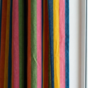 May include: A vibrant curtain featuring vertical stripes in orange, pink, green, yellow, and blue. The fabric has a textured appearance, possibly linen or cotton. The curtain hangs in front of a window.