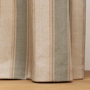 Op de afbeelding: Close-up van een beige en groen gestreepte stof. De verticale strepen wisselen af tussen bredere beige secties en smallere strepen van groen en lichtbruin. De stof lijkt een zware weving te zijn, mogelijk linnen of katoen.