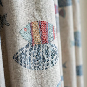 May include: Beige fabric curtain with embroidered fish and starfish designs. The fish are in shades of blue, red, and yellow, with a detailed stitched pattern. The starfish and other shapes are in blue. The fabric has a textured appearance.